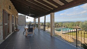 Un patio techado con una mesa y sillas de comedor, un muro de piedra y vistas a una piscina y el paisaje.