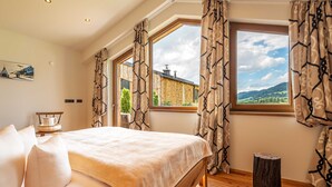 Une chambre avec un grand lit, deux fenêtres avec des rideaux, un plancher en bois et un petit tabouret.