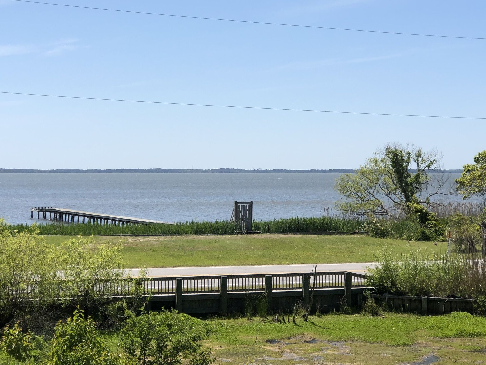 A lakeside view with a wooden dock extending into the water, a grassy area with a bench, and a clear sky.