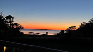 A wooden deck with railings and lights, overlooking a sunset over water with a pier extending into the distance. - Beach House Rental Duck, NC (Duck)