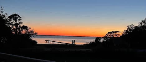 Une terrasse en bois, dotée de rampes et d’éclairages, qui offre une vue sur un coucher de soleil se reflétant sur l’eau, avec une jetée s’étendant au loin.