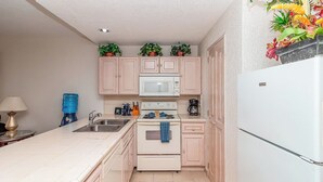 Cocina compacta con refrigerador blanco, microondas, estufa y pileta. Hay alacenas sobre la estufa y un dispensador de agua en la encimera.