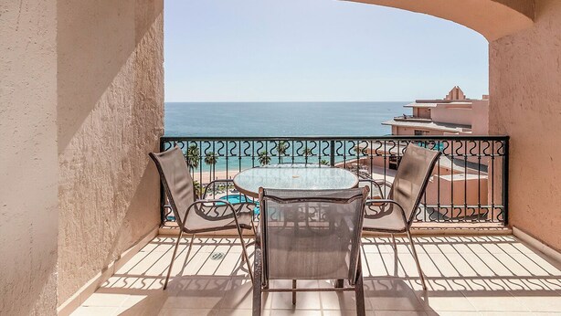 Un balcón con mesa y sillas, con vistas a una piscina y edificios, y una vista despejada del mar.