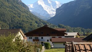 A mountain house with a sloped roof, surrounded by lush greenery and a snow-capped peak in the background.