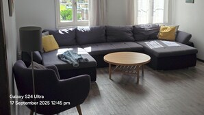 A modern living room with a grey sectional sofa, a round wooden coffee table, and a dark armchair. A clock and a painting are visible on the walls.