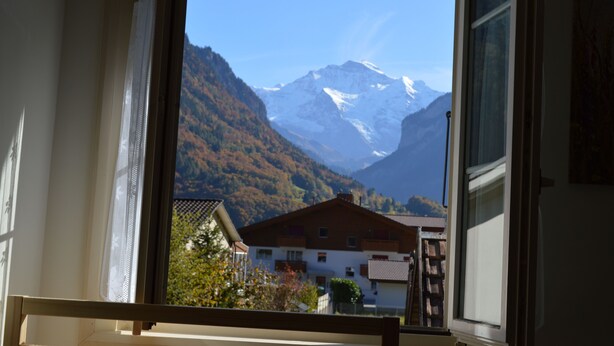 A view from a window overlooking a mountain with snow-capped peaks and a building with a sloped roof.