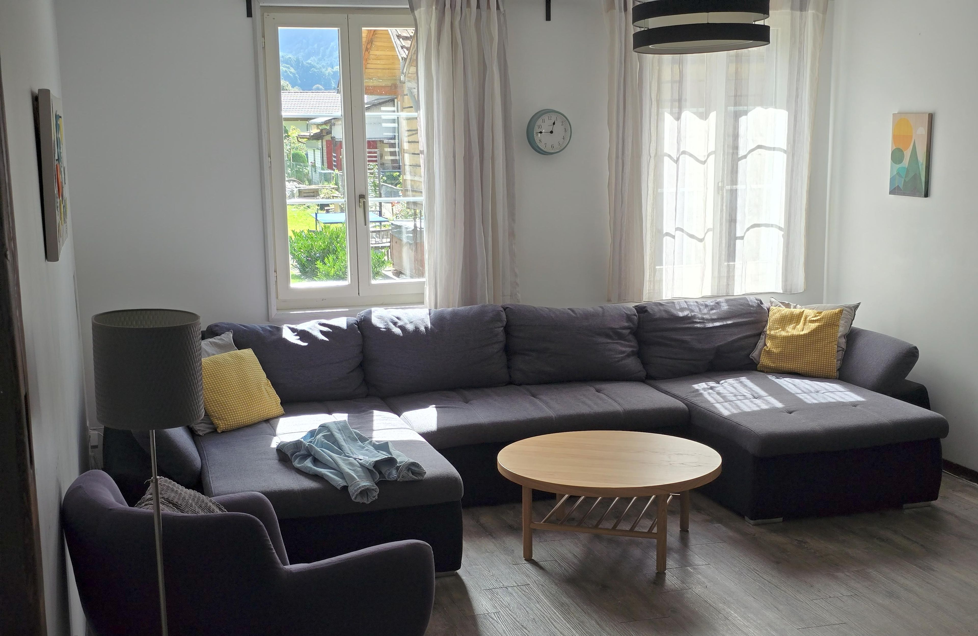 A modern living room with a grey sectional sofa, a round wooden coffee table, and a dark armchair. A clock and a painting are visible on the walls.