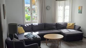 A modern living room with a grey sectional sofa, a round wooden coffee table, and a dark armchair. A clock and a painting are visible on the walls.