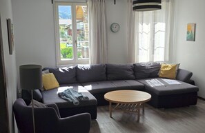A modern living room with a grey sectional sofa, a round wooden coffee table, and a dark armchair. A clock and a painting are visible on the walls.