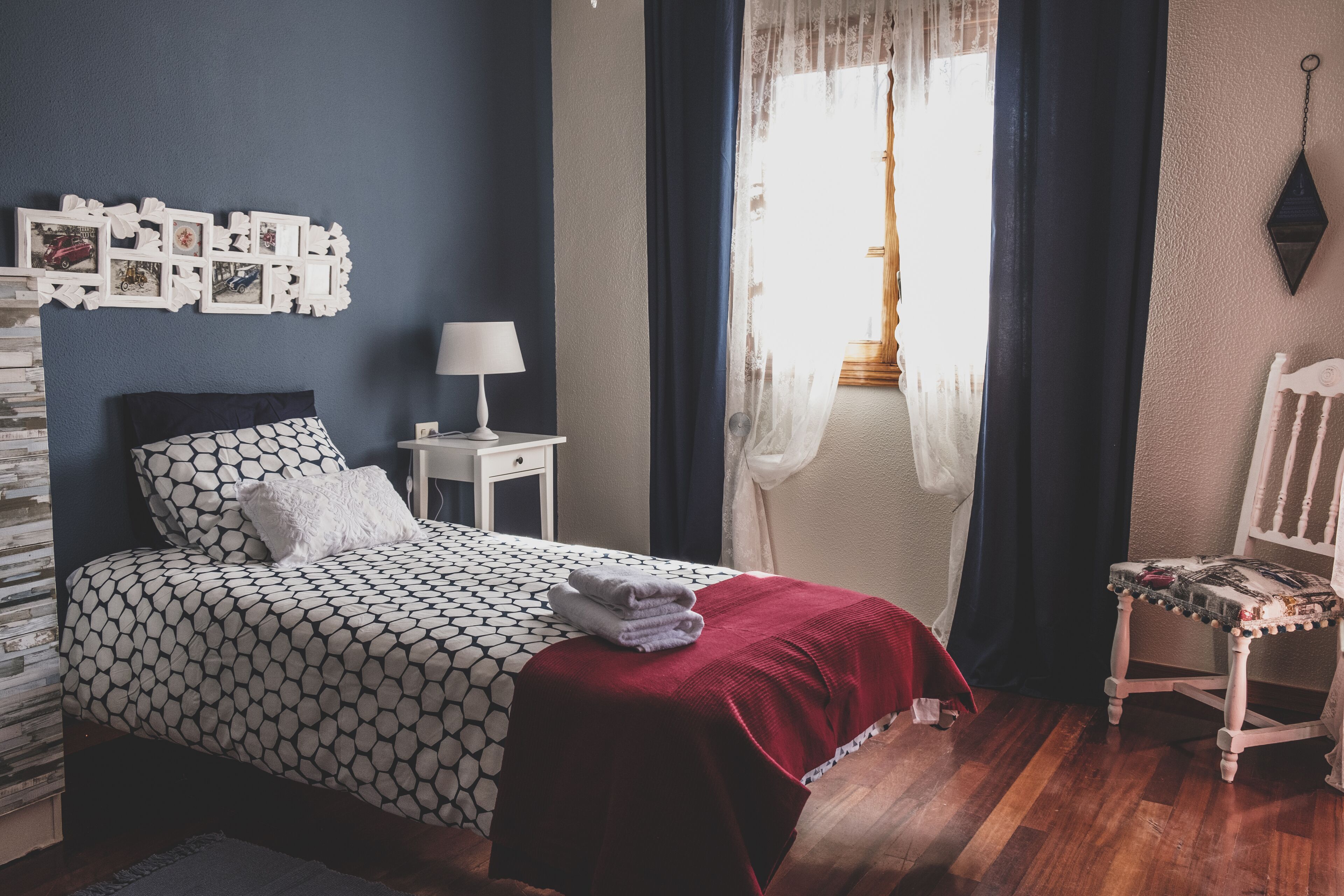 A neatly arranged bedroom with a bed, a bedside table with a lamp, a chair, and a window with curtains.