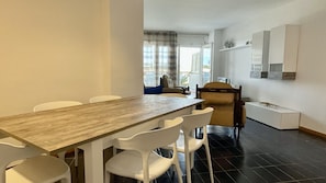 A dining area with a long wooden table and white chairs, a small kitchen area with a white cabinet, and a view through a window with curtains.