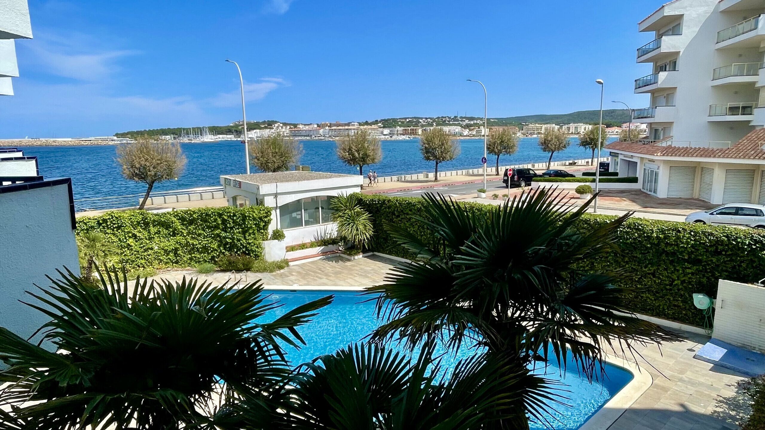 A pool area with a view of the sea, a modern building, and a road with pedestrians.