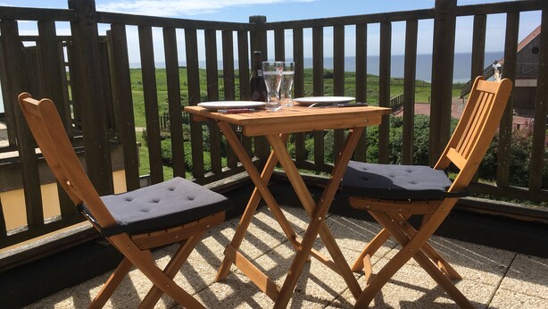 Een houten tafel en twee stoelen op een balkon, met uitzicht op de zee.