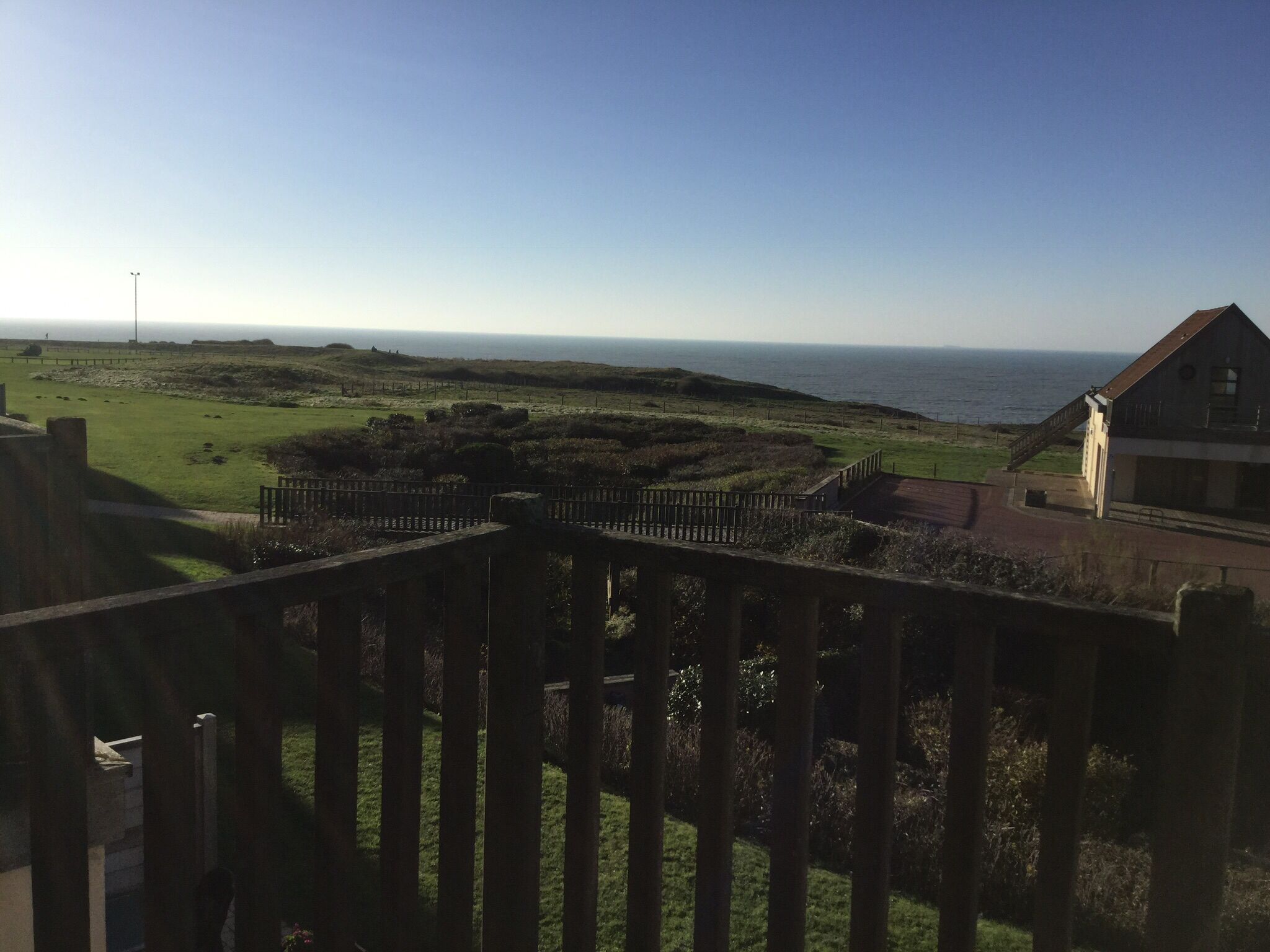 A coastal view from a balcony with a wooden railing, grassy fields, and a building with a red roof in the foreground.