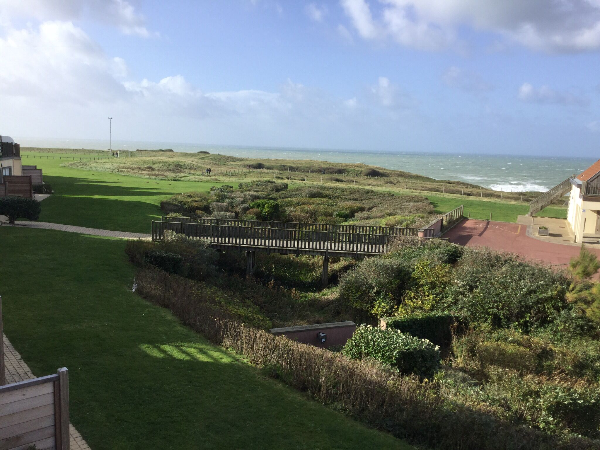 A coastal view with a wooden bridge, grassy areas, and a building with a red roof.