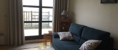 Ein Wohnzimmer mit einem blauen Sofa, zwei Kissen, einem kleinen Holztisch, einer Lampe und Blick durch ein großes Fenster.