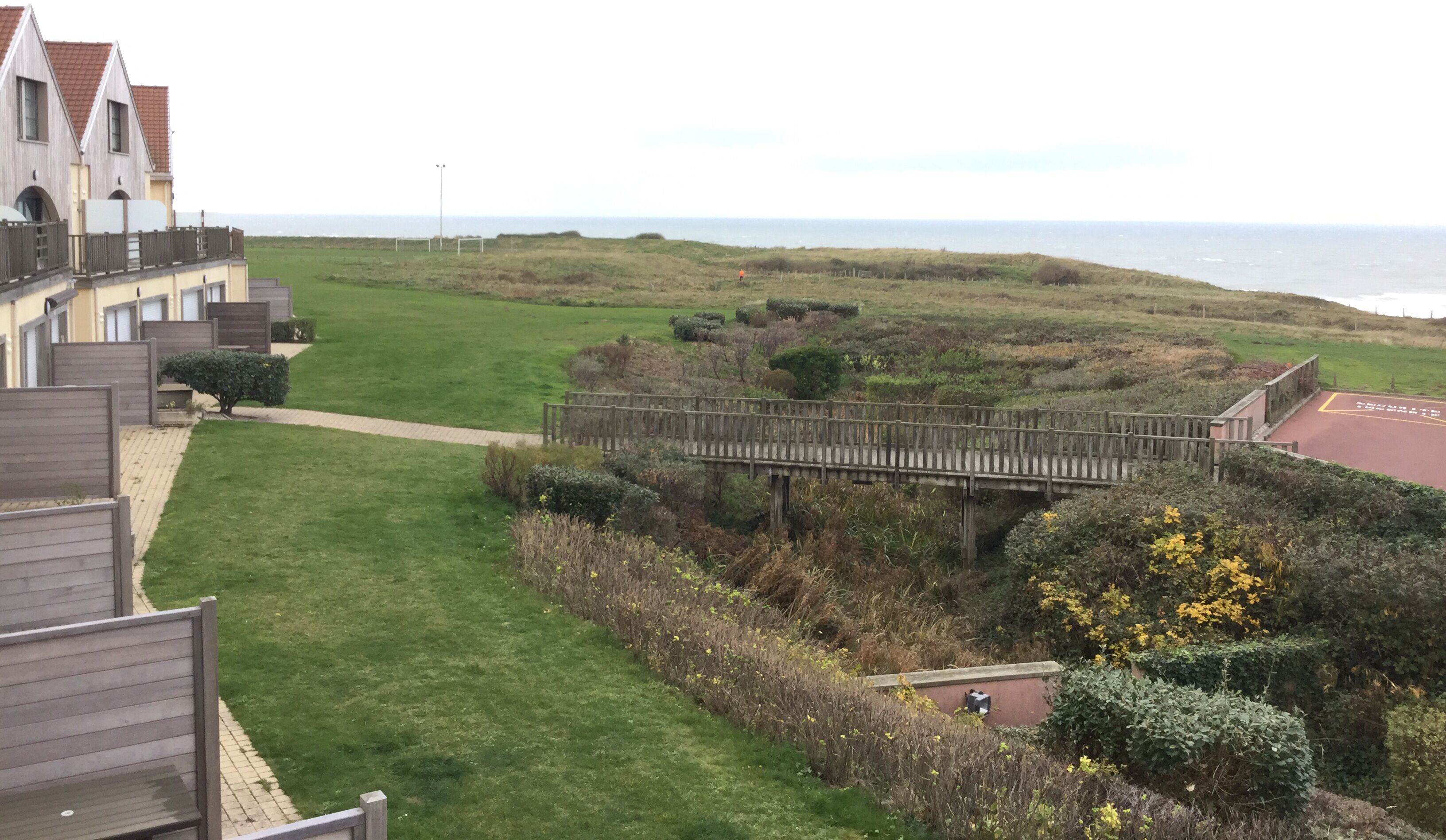 A coastal residential area with a wooden bridge, grassy fields, and a view of the sea.