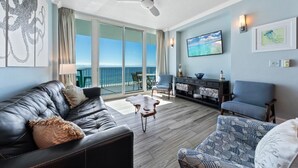 A spacious living room with a leather sofa, patterned armchair, and a coffee table. A ceiling fan is visible, and there is a large window offering a view of the ocean. - Lighthouse 714 (Gulf Shores)