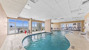An indoor swimming pool with clear views of the ocean through large windows, a fitness area with treadmills, and a well-lit ceiling. - Lighthouse 714 (Gulf Shores)