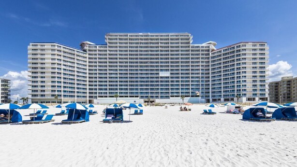 A large hotel with multiple floors and balconies, a sandy beach in front, and several beach umbrellas. - Lighthouse 714 (Gulf Shores)