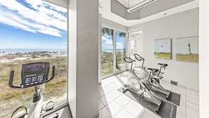 A modern gym with a stationary bike and elliptical, large windows with a view of the beach and palm trees. - Lighthouse 714 (Gulf Shores)