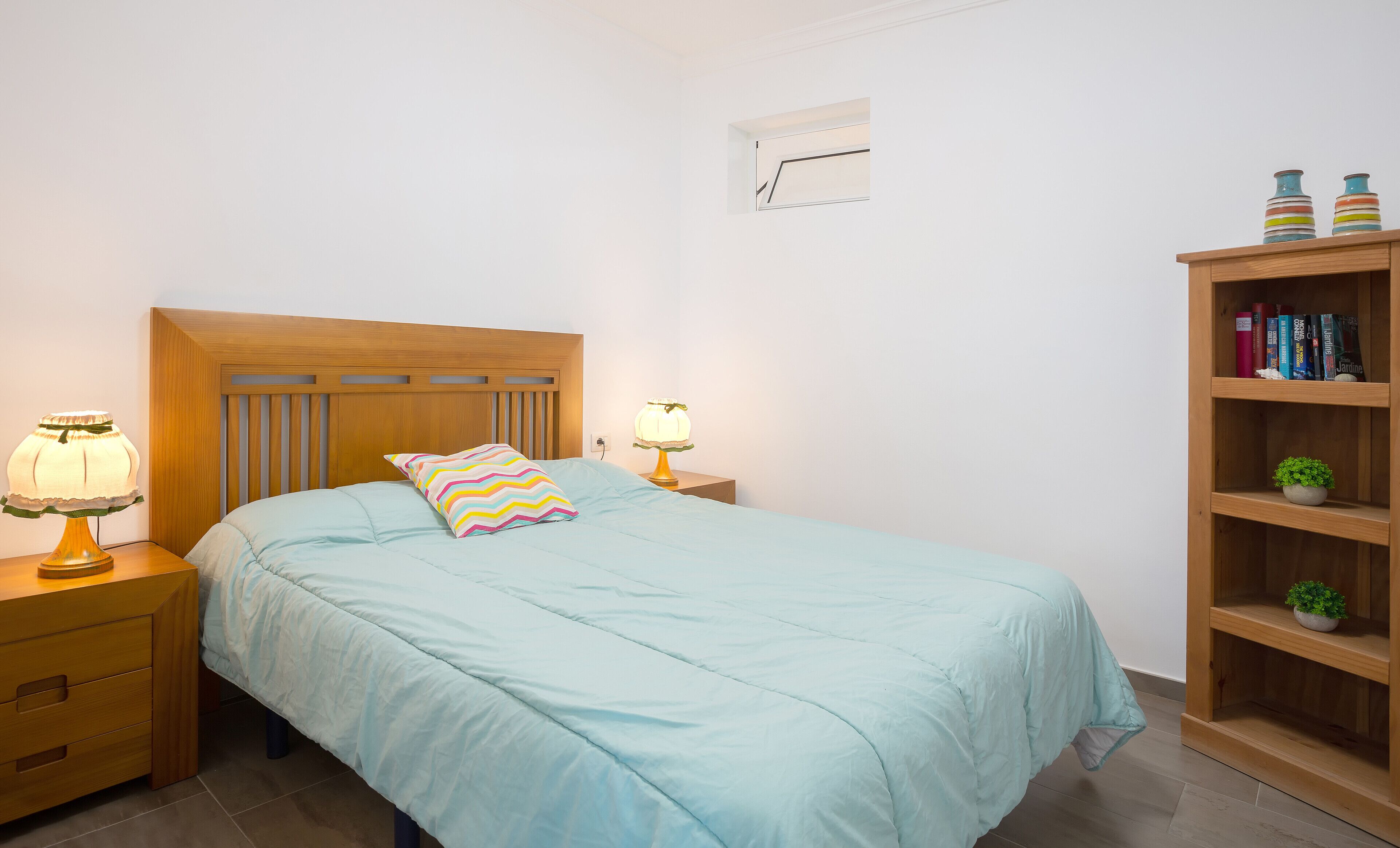 Un dormitorio con una cama, dos mesitas de noche con lámparas, un cabecero de madera, un reloj de pared y una estantería con libros y una pequeña planta.