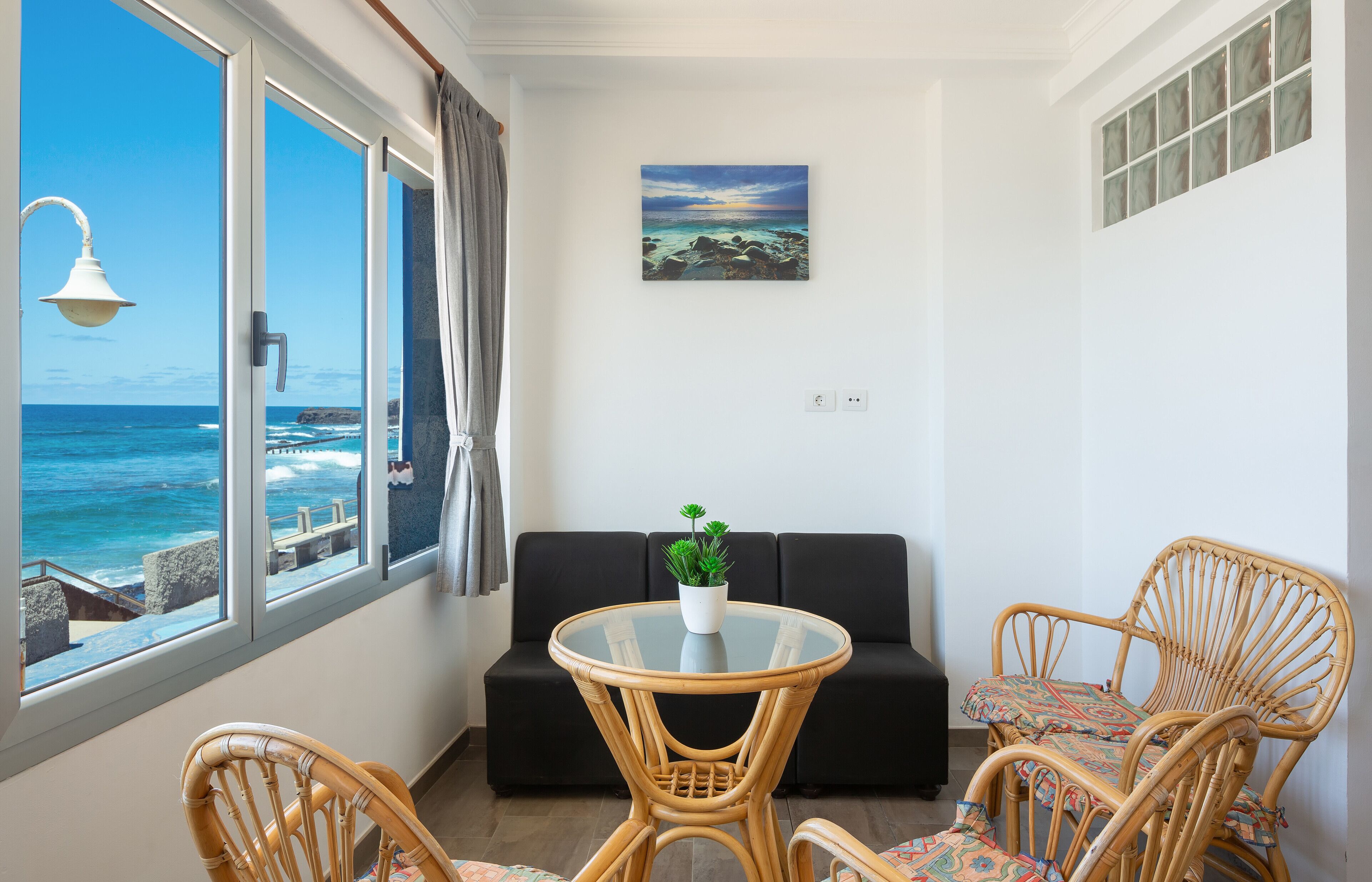 Une pièce avec une table en verre, un canapé noir, des chaises en osier, une plante en pot et un tableau.