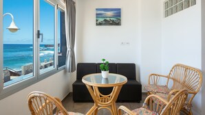 Een kamer met een glazen tafel, een zwarte bank, rieten stoelen, een potplant en een schilderij.