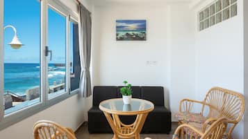 Une pièce avec une table en verre, un canapé noir, des chaises en osier, une plante en pot et un tableau.