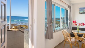 Een balkon met uitzicht op de oceaan, een rieten stoel, een klein tafeltje en een vaas met bloemen.