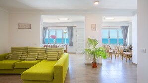 Een lichte woonkamer met een groene bank, een eethoek met rieten stoelen en uitzicht op zee door grote ramen.