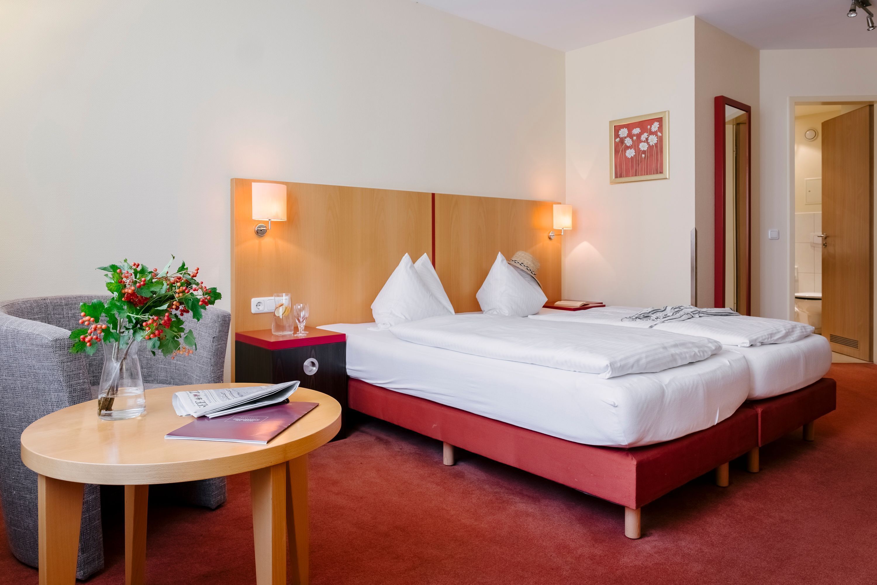 A hotel room with a bed, bedside tables, a small table with a vase and a remote control, and a door leading to a bathroom.