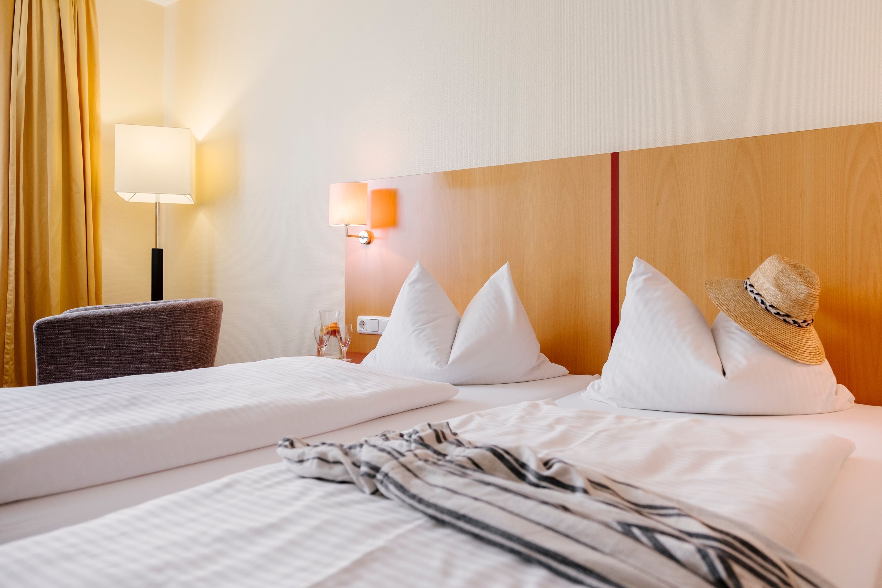 A hotel room with a bed, a chair, a lamp, and a hat on the bed.