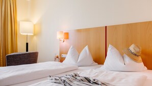 Una habitació d'hotel amb un llit, una cadira, una làmpada i un barret sobre el llit.
