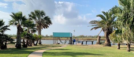 A grassy area with a concrete path leading to a dock, palm trees, and a blue structure by the water.