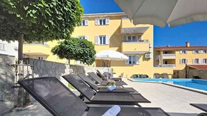 Uma área à beira da piscina com espreguiçadeiras, piscina e um edifício com varandas ao fundo.