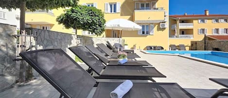 A poolside area with sun loungers, a pool, and a building with balconies in the background.