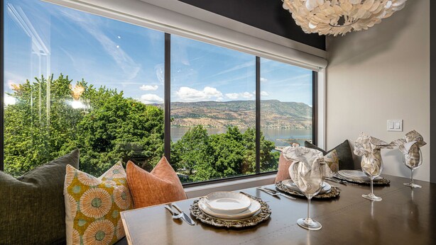 A dining area with a table set for two, a large window offering a scenic view, and a decorative chandelier. - Designer Penthouse - Downtown Waterfront! (Kelowna)