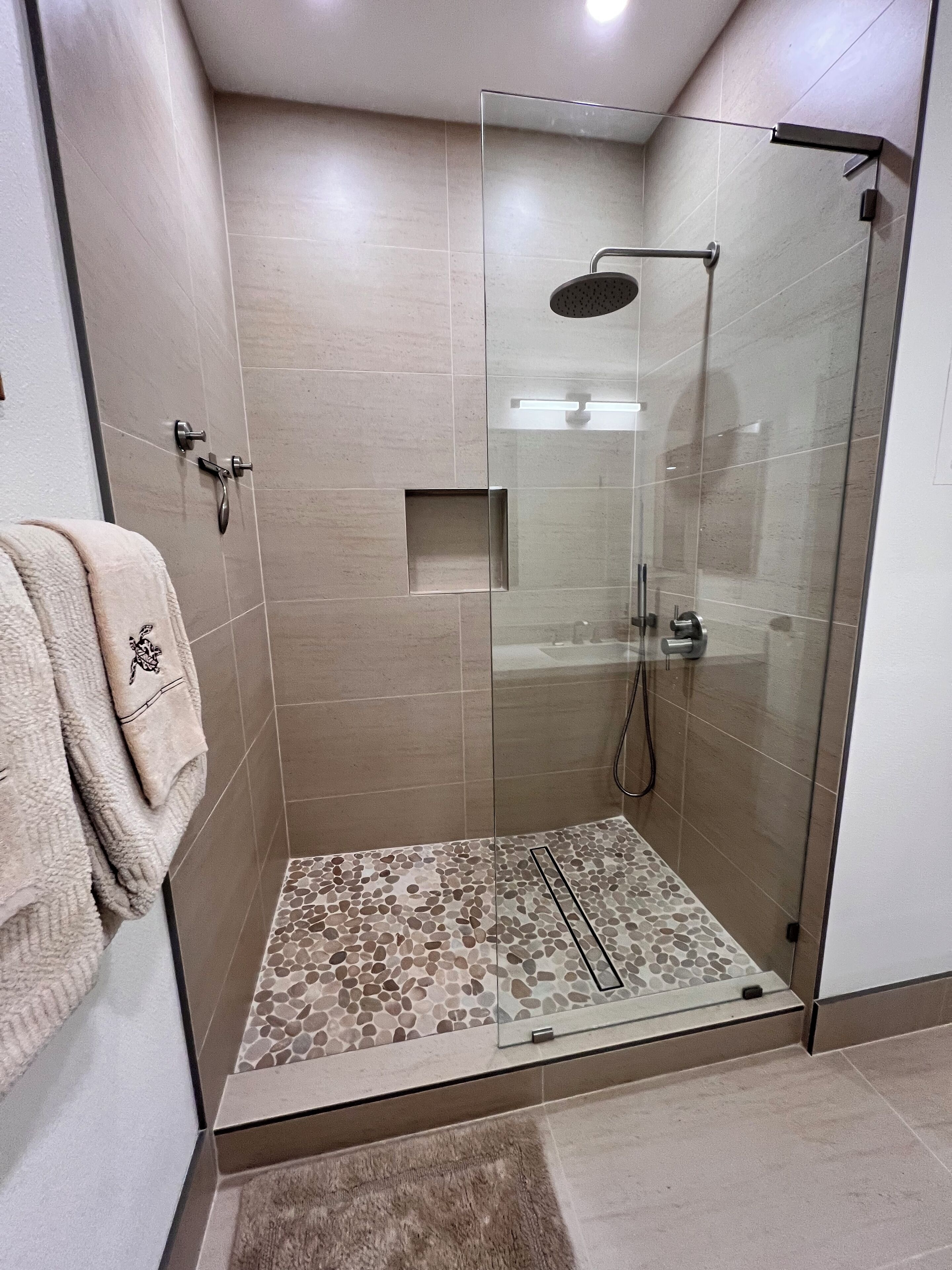 Une douche fermée par des parois en verre, dotée d’un pommeau de douche à effet pluie, d’une petite étagère carrée et d’un sol texturé.