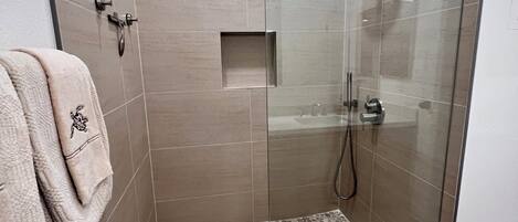 Une douche fermée par des parois en verre, dotée d’un pommeau de douche à effet pluie, d’une petite étagère carrée et d’un sol texturé.
