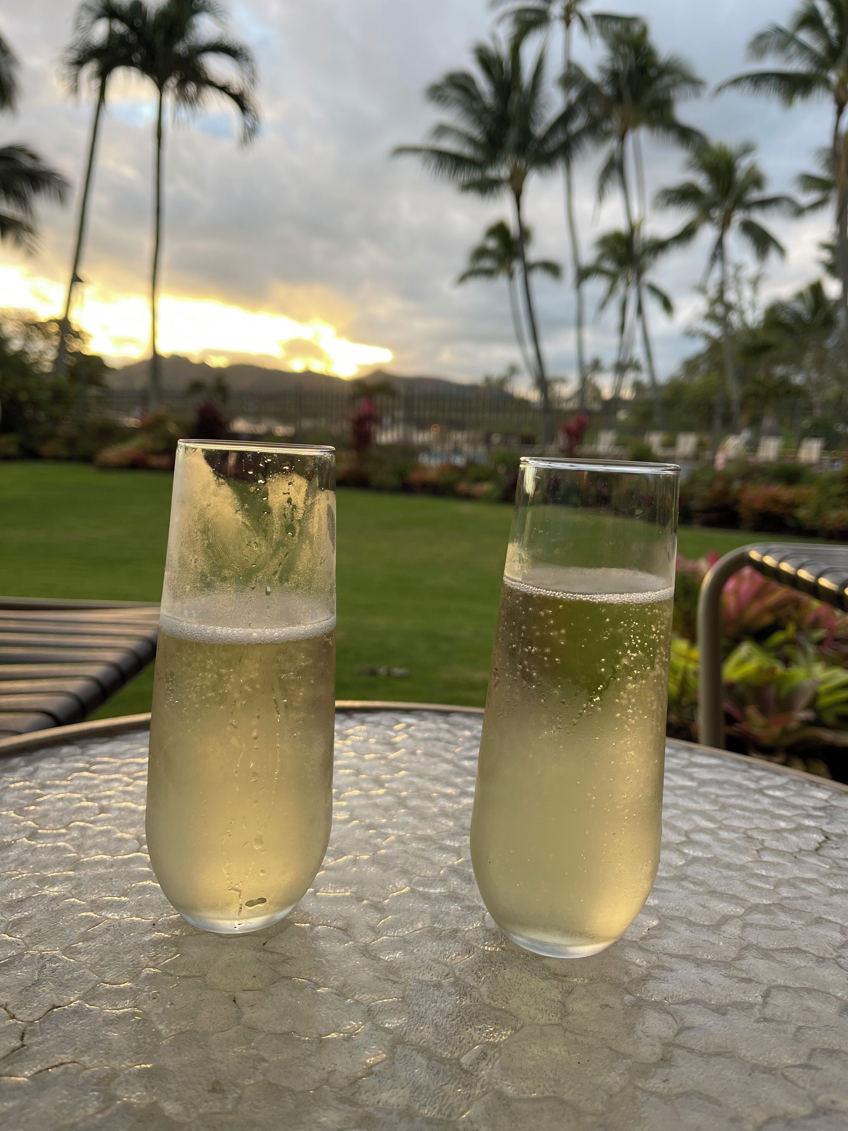 Dos vasos de una bebida de color claro sobre una mesa exterior con textura, con vistas a palmeras y un atardecer al fondo.