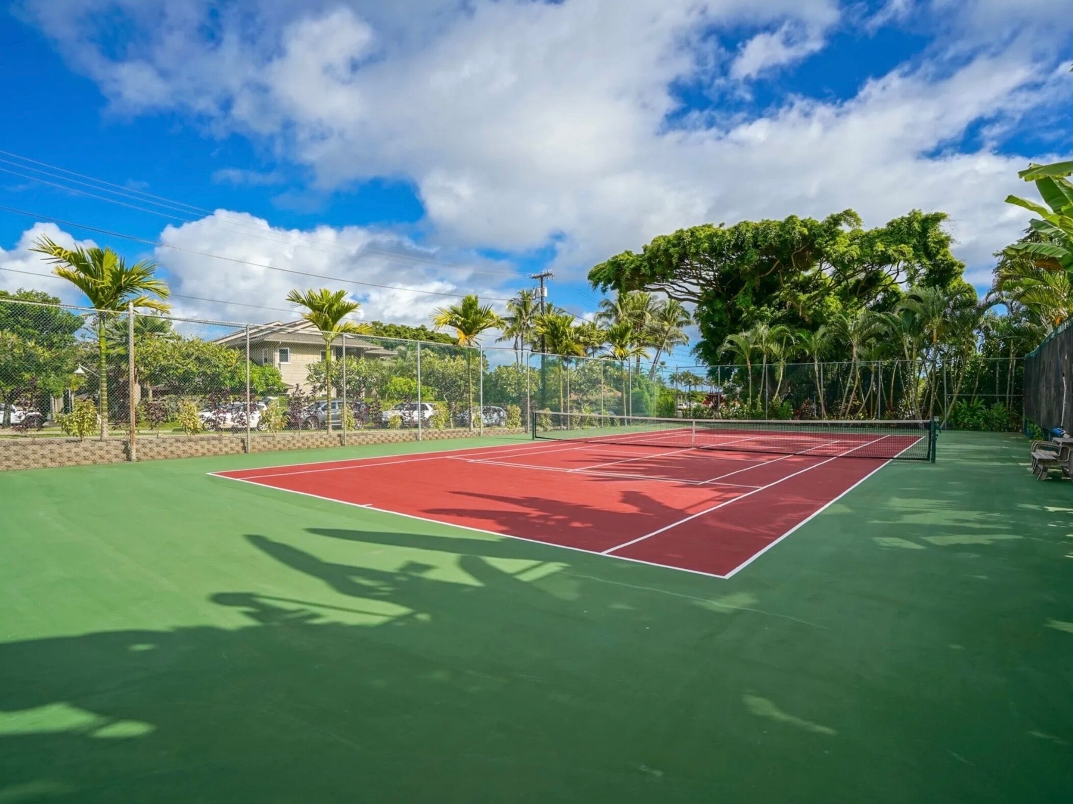 An outdoor tennis court with a red playing surface, surrounded by greenery and a clear sky.