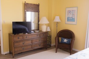 A room with a wooden dresser, a television, two lamps, a wicker chair, and a framed picture on the wall.