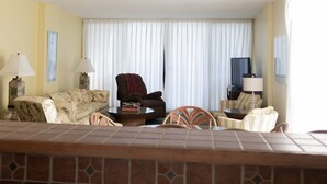 Habitación de hotel con sofá, sillones, una mesa de centro, televisión y un ventanal con cortinas.