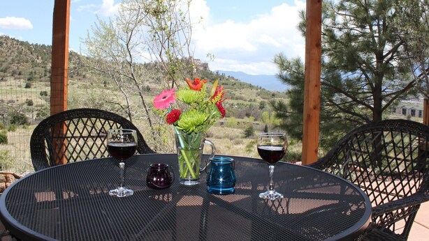 Una mesa redonda con cubierta metálica, rodeada de sillas de mimbre negro, ubicada al aire libre con vistas a un paisaje montañoso y árboles al fondo.