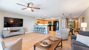 A modern living room with a TV, sofa, and coffee table, connected to a kitchen with bar stools. - Spacious, Luxury, Beach Condo with Pool (Indian Rocks Beach)