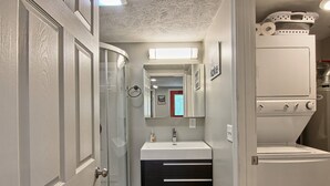 Un baño pequeño con un lavamanos blanco, cajones negros, cabina de ducha de vidrio y una lavadora blanca.
