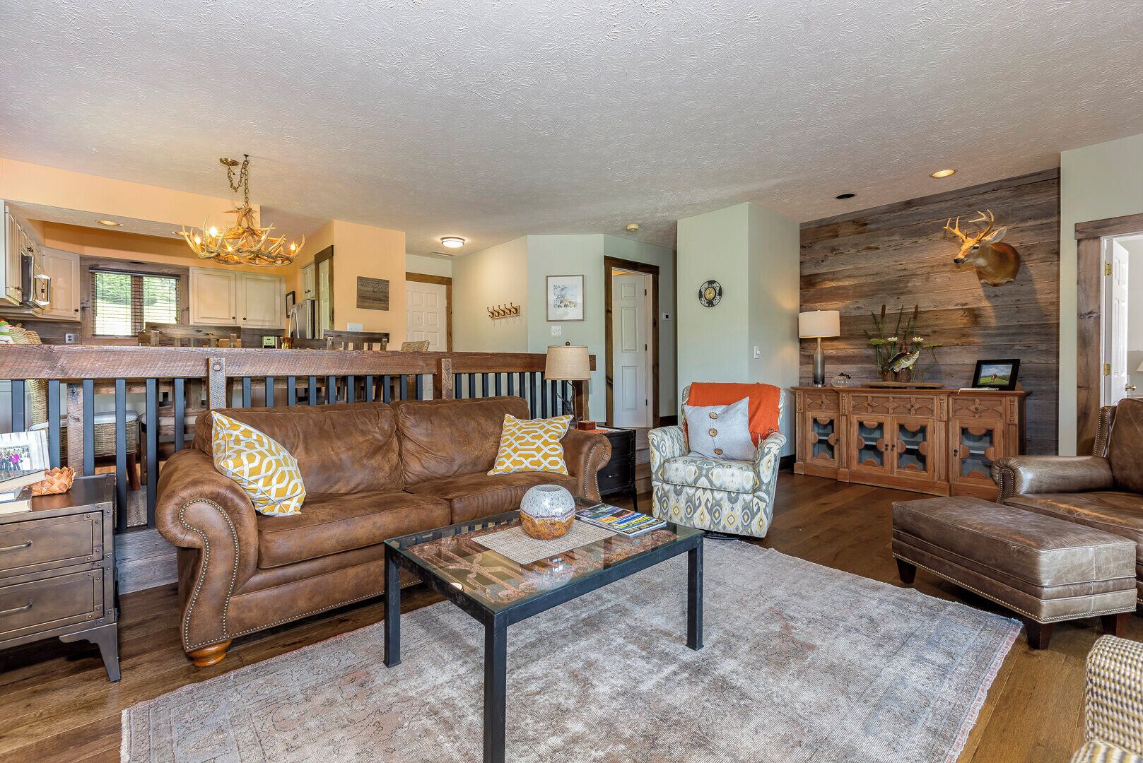 A spacious living room with a brown leather sofa, patterned armchair, and ottoman. A glass coffee table is in the center.