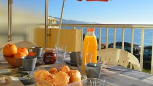 Une table dressée avec des aliments pour le petit-déjeuner, notamment des oranges, une bouteille de jus d’orange et un pot de confiture, sur un balcon donnant sur la mer.
