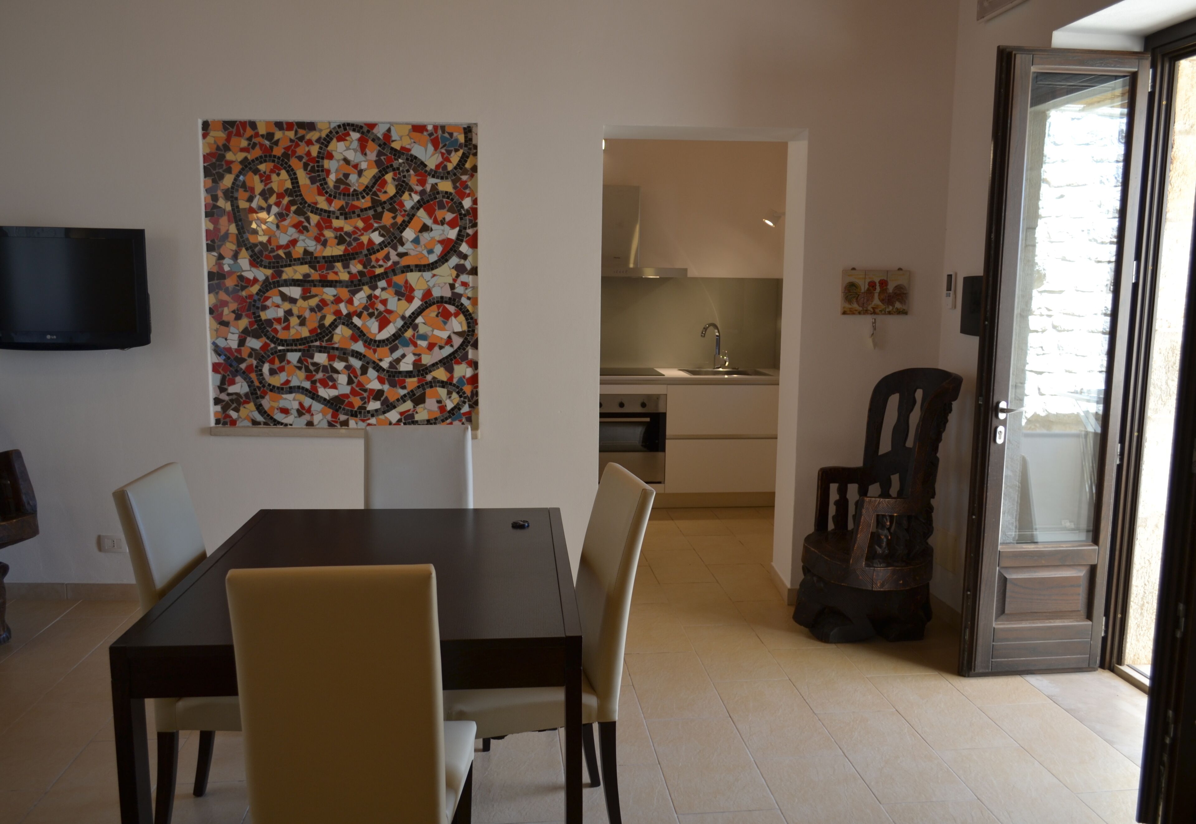 A dining area with a table and chairs, a kitchen in the background, and a television mounted on the wall.